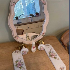 May include: A set of decorative ceramic door knobs and plates with floral designs. The knobs are gold and the plates are white with pink and green flowers. The set also includes a small ceramic hook with a floral design.