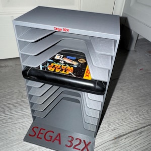 Pode incluir: Um suporte cinza impresso em 3D para cartuchos de jogos Sega 32X. O suporte possui vários slots para cartuchos de jogos e apresenta o texto "Sega 32X" em vermelho. Um cartucho de jogo Star Wars é visível em um dos slots.