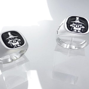 May include: Two silver rings featuring a black enamel design of a cartoon character wearing a hat and holding a sign with the number "II" on it. The rings are displayed on stands.