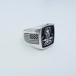 May include: A silver ring with a black enamel inlay featuring an American flag on one side and a military insignia on the other.