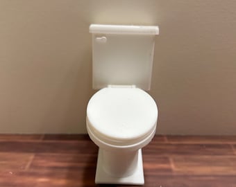 May include: A miniature white toilet with a closed lid, set against a neutral wall and a wood-grain floor. The toilet has a rectangular base and a rounded bowl. The tank has a lever on the side.
