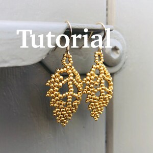 May include: Gold beaded earrings in a leaf shape with a delicate design. The earrings are hanging from a hook.