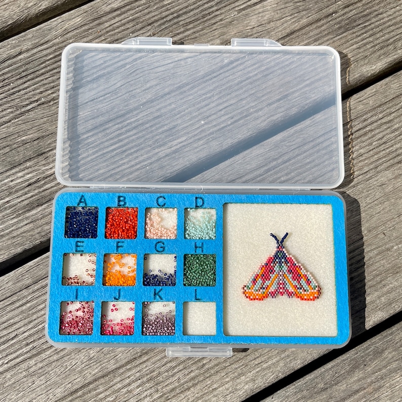 May include: A blue plastic storage case with a clear lid. The case has 12 compartments labeled A through L. Each compartment contains small beads of different colors. A beaded butterfly design is displayed in the center compartment.