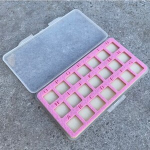 May include: A clear plastic storage box with a pink foam insert. The insert has 26 compartments labeled with letters from A to U.