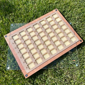 Könnte beinhalten: Ein Holzbrett mit einem Raster aus Quadraten, die jeweils einen Buchstaben des Alphabets enthalten. Das Brett ist mit einer durchsichtigen Acrylglasscheibe bedeckt. Die Buchstaben sind in brauner Schrift gedruckt.