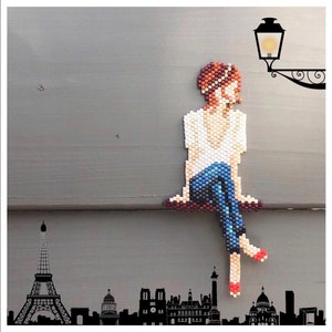 May include: A beaded artwork depicting a person sitting with their legs crossed, wearing a white shirt and blue jeans. The person has brown hair and is wearing red shoes. The artwork is against a grey background with a black silhouette of a city skyline at the bottom.
