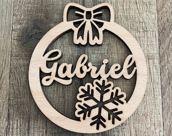 May include: A wooden Christmas bauble with a bow detail at the top. The name "Gabriel" is cut out in cursive script, with a snowflake design at the bottom. The bauble is made from light-coloured wood.