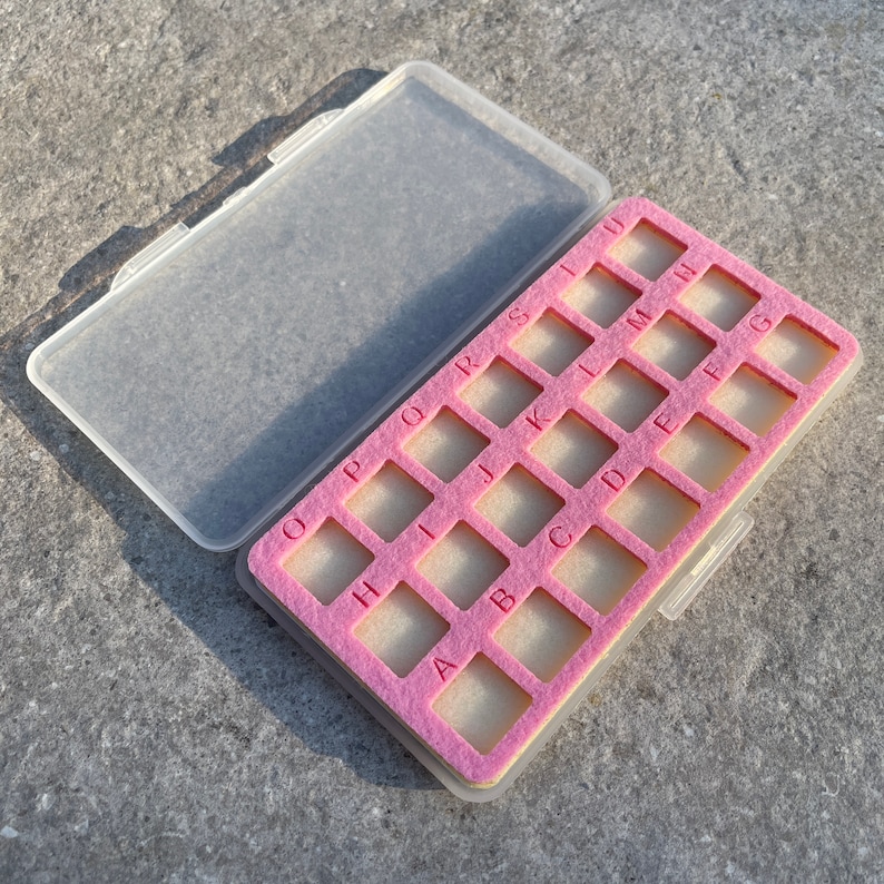 May include: A clear plastic storage box with a pink felt interior. The box has 19 compartments labeled with letters from A to S.