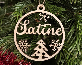 May include: A wooden Christmas bauble with the name "Satine" cut out in a cursive font. The circular ornament features snowflakes and a Christmas tree design. It hangs from a natural twine string.