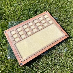 Könnte beinhalten: Ein Holzbrett mit einem Raster von Fächern, die mit Buchstaben des Alphabets beschriftet sind. Das Brett ist mit einer durchsichtigen Acrylglasscheibe abgedeckt.