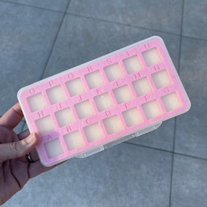 May include: A clear plastic pill box with a pink interior and a felt lining. The box is labeled with letters from A to U.