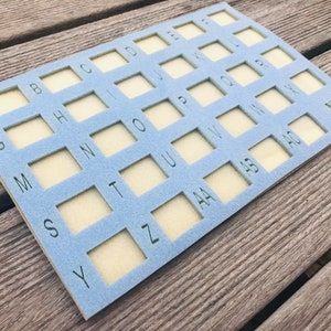 May include: A light blue felt board with a grid of squares, each labeled with a letter of the alphabet. The board is designed for organizing and storing small items.