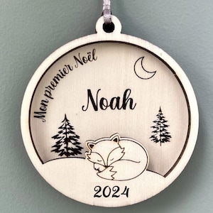 Peut inclure: Une décoration de Noël en bois avec un renard endormi, deux sapins, une lune croissante et le texte "Mon premier Noël" et "Noah" avec l'année "2024" en dessous.