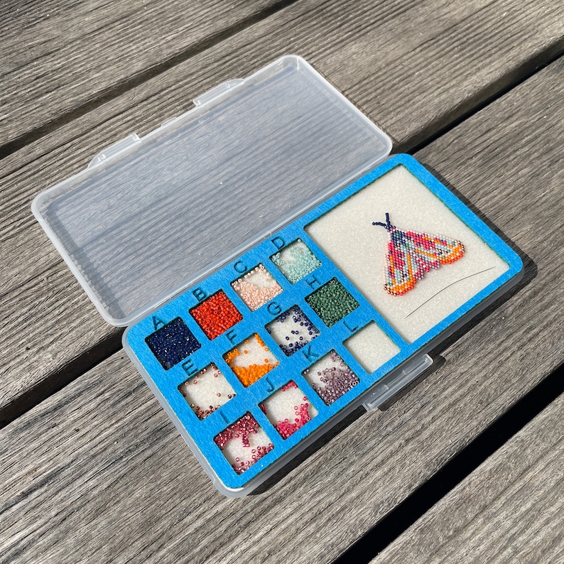 May include: A blue plastic storage case with a clear lid, containing small containers of colorful beads labeled A through L. A beaded moth design is displayed on the case.