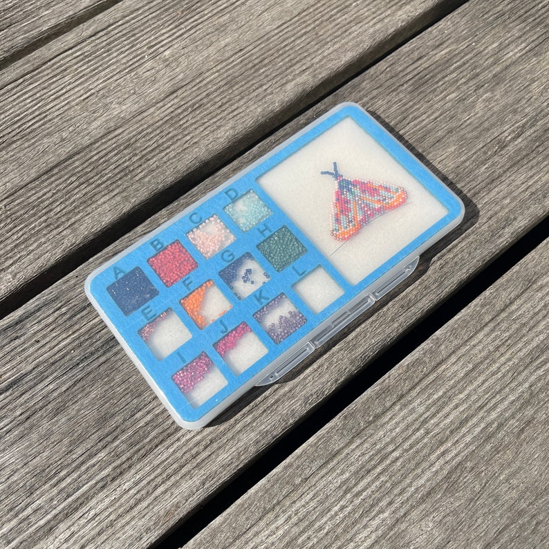 May include: A blue plastic bead organizer with a clear lid. The organizer has 12 compartments labeled with letters A through L. Each compartment contains a variety of colorful beads. A beaded butterfly design is visible in the top compartment.