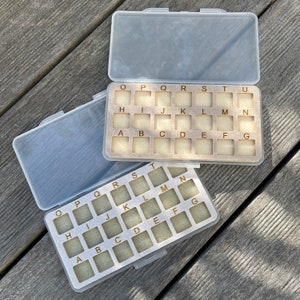 May include: A clear plastic storage case with two compartments. Each compartment has a grid of 21 small squares labeled with letters of the alphabet. The top compartment has the letters O, P, Q, R, S, T, U, H, I, J, K, L, M, N, A, B, C, D, E, F, and G. The bottom compartment has the letters O, P, Q, R, S, T, H, I, J, K, L, M, N, A, B, C, D, E, F, and G.