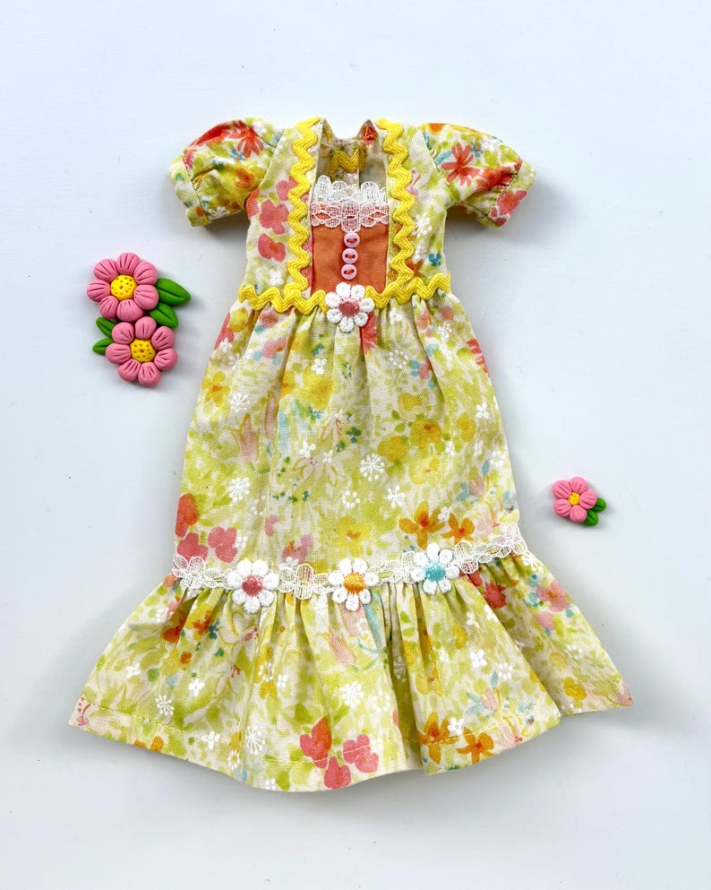 May include: A yellow and white floral print dress with a lace trim and orange and yellow accents. The dress has a ruffled hem and is decorated with small flowers.