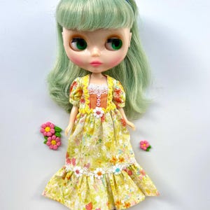 May include: A green haired doll wearing a yellow floral dress with a lace trim and orange accents. The doll has green eyes and is wearing a blue headband. There are pink flowers on the white background.
