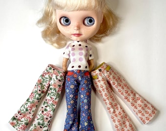 May include: A doll with blonde hair, blue eyes, and a white shirt with pink polka dots. The doll is wearing three pairs of flared trousers with floral patterns. Pink sandals are on the doll's feet. A blue headband is on the doll's head.