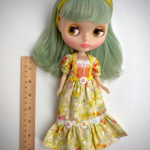 May include: A green haired doll wearing a yellow and orange floral dress with white lace trim and a yellow headband. The doll has brown eyes and pink cheeks.