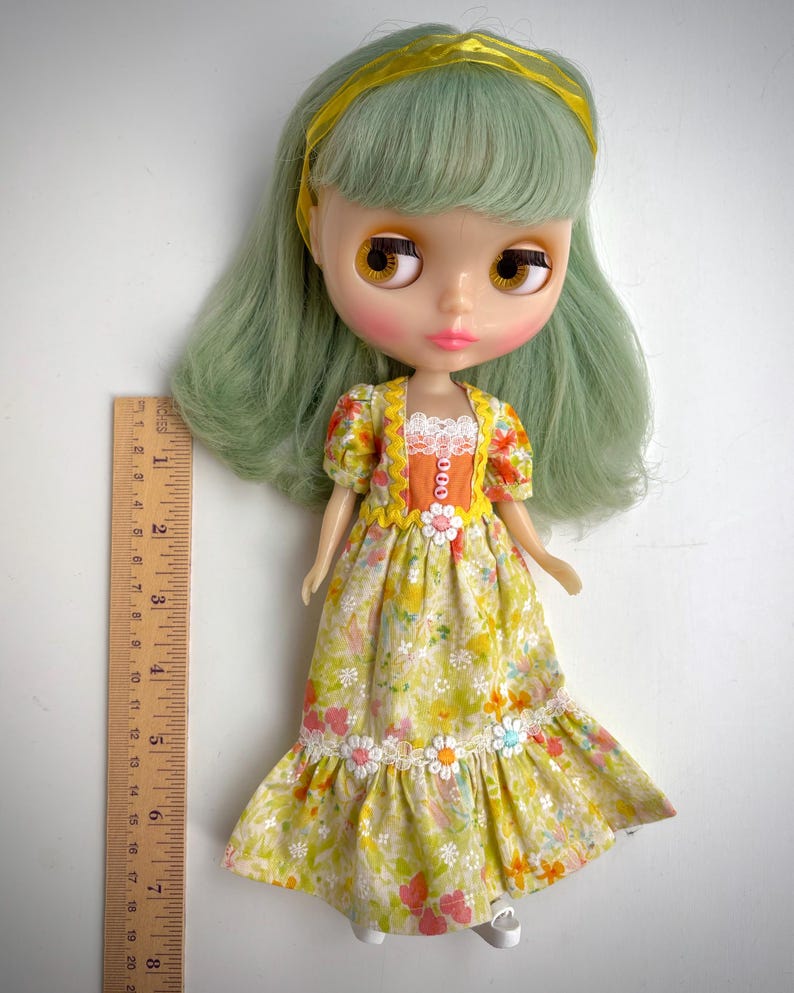May include: A green haired doll wearing a yellow and orange floral dress with white lace trim and a yellow headband. The doll has brown eyes and pink cheeks.