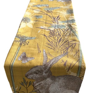 May include: A yellow table runner featuring a brown rabbit, green leaves, and a butterfly design.