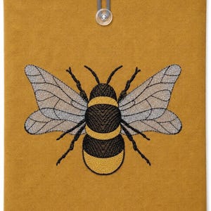 Könnte beinhalten: Eine rechteckige Stoffhülle in Senfgelb mit einer mittig aufgestickten Hummel, die schwarze und gelbe Streifen sowie durchscheinende graue Flügel aufweist. Am oberen Rand befindet sich eine graue elastische Schlaufe mit einem Knopf.