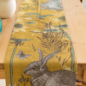 May include: A rectangular table runner in a mustard yellow hue featuring a repeating print of grey rabbits, butterflies, and botanical elements in shades of teal and dark grey. The fabric has a visible weave texture and a vertical orientation.