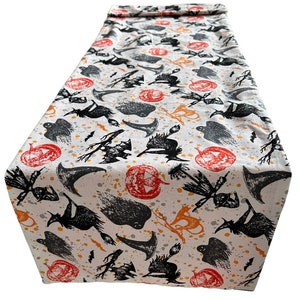 May include: A rectangular table runner featuring a cream-coloured fabric base with a repeating Halloween-themed print. The pattern includes black silhouettes of witches on broomsticks, pointed hats, bats, ghosts, and orange jack-o'-lanterns.