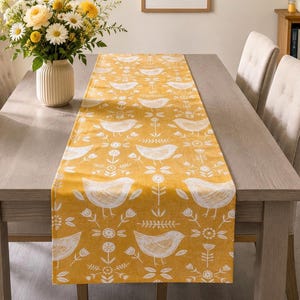 May include: A rectangular table runner in a vibrant mustard yellow colour featuring a repeating white pattern of stylised birds, small flowers, and leafy sprigs. The fabric has a textured appearance and is laid flat across a wooden dining table.