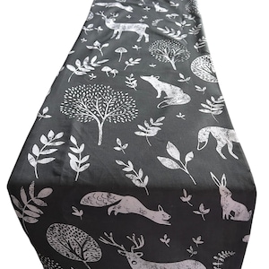 May include: A dark gray table runner with a white woodland pattern. The design includes deer, foxes, rabbits, trees, and foliage. The runner is folded at the top.