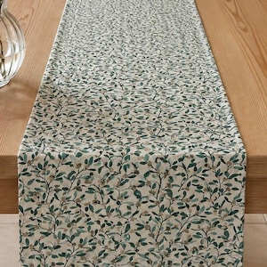 May include: A rectangular table runner featuring a repeating botanical pattern of dark green and muted brown leaves on a light cream background. The fabric is draped lengthwise across a wooden table surface, displaying a dense, intricate foliage design.