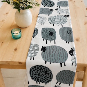 May include: A white table runner featuring a repeating pattern of stylized sheep in various shades of gray and black. Each sheep has a unique texture, including dots, swirls, and dashes, printed across the fabric surface in a vertical orientation.