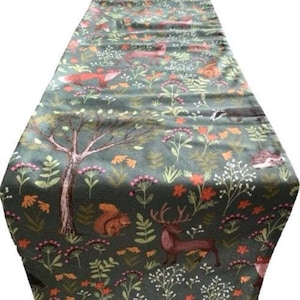 May include: A green table runner with a woodland pattern featuring foxes, deer, squirrels, and other forest animals. The runner has a dark green background with a variety of colourful flowers and leaves.