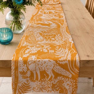 May include: A rectangular table runner featuring a mustard yellow background with a white repeating pattern of woodland animals including foxes, owls, squirrels, and rabbits, interspersed with detailed illustrations of ferns and forest foliage.