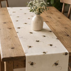 May include: A cream-coloured table runner featuring embroidered bees. A small, beige vase holds a bouquet of white flowers. The runner is on a wooden table, creating a rustic, farmhouse aesthetic. The table is made of wood.