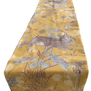 May include: A yellow table runner with a repeating pattern of brown rabbits, green grass, and blue flowers.