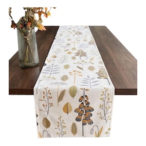 May include: A white table runner featuring a repeating botanical pattern of leaves, stems, and flowers in shades of mustard yellow, brown, and grey. The runner is laid out flat on a dark wood surface with a glass vase of dried foliage nearby.