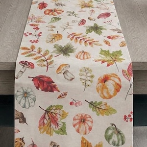 May include: A rectangular table runner featuring a cream-coloured fabric base printed with a repeating pattern of autumn-themed illustrations including orange and green pumpkins, red and yellow maple leaves, brown acorns, and red-capped mushrooms.