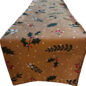 May include: A tan table runner featuring a repeating design of green holly leaves, red berries, and white dots. The runner is folded over at the top, displaying the pattern. Ideal for festive holiday table settings.