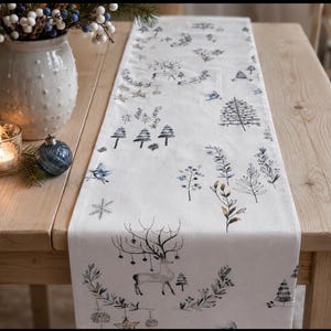 May include: A white table runner with a winter design, featuring deer, trees, and botanical elements in blue and grey. The runner is on a light wooden table, with a vase of berries, a candle, and a blue bauble.