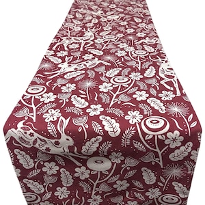 May include: A burgundy table runner with a repeating white pattern of rabbits, flowers, and foliage. The runner is rectangular and appears to be made of fabric. The design is a whimsical, nature-inspired print.