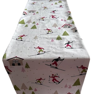 May include: White table runner with a winter ski scene. The runner features skiers in red and black outfits, green trees, and pink and red houses. The design is repeated across the fabric.