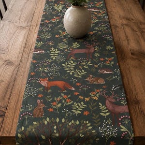May include: A dark green table runner featuring a repeating pattern of woodland animals including deer, foxes, rabbits, hedgehogs, and badgers, interspersed with colourful wildflowers, green foliage, and trees, topped with a beige ceramic vase.