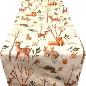 May include: A white table runner with a woodland pattern featuring deer, foxes, rabbits, birds, and trees. The pattern is in shades of brown, orange, and green.