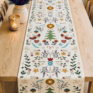 May include: A long rectangular table runner featuring a repeating pattern of green Christmas trees, gold stars, red hearts, blue ornaments, and stylised floral foliage on a cream-coloured fabric background, centred on a light wood dining table.