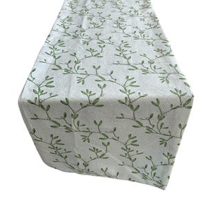 May include: A light-colored table runner with a repeating pattern of green leaves and small white dots. The fabric is folded at the top, showcasing the design. This decorative runner is suitable for a dining table or other surfaces.