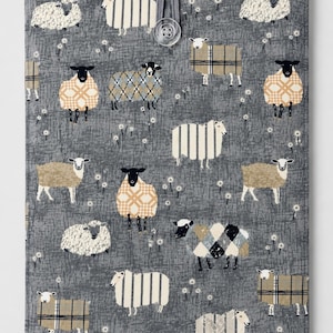 May include: A rectangular fabric cover featuring a dark gray background with a repeating pattern of sheep in various styles, including solid, striped, and plaid designs. Small white dandelion-like flowers are scattered throughout. A clear button closure.