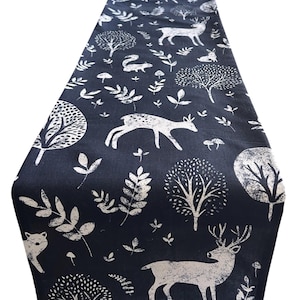 May include: A dark blue table runner featuring a white forest design. The pattern includes deer, trees, foxes, and leaves. The fabric runner is suitable for a dining table or other surface.