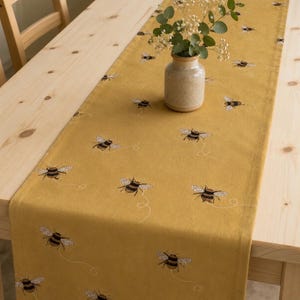 May include: Mustard yellow table runner featuring embroidered bees. A small vase with greenery sits on the runner, which is placed on a light wood table. The runner adds a touch of nature to the dining area. The table is made of light wood.